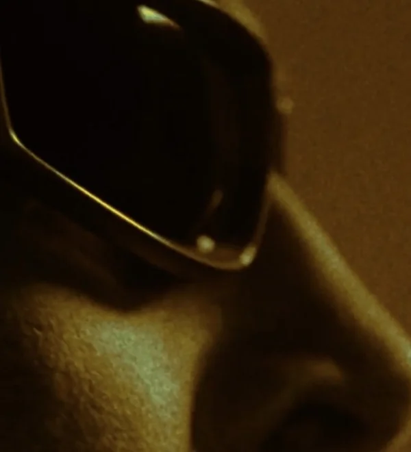 Close-up of a person wearing dark sunglasses in low light, showing the nose and cheek area with amber lighting.