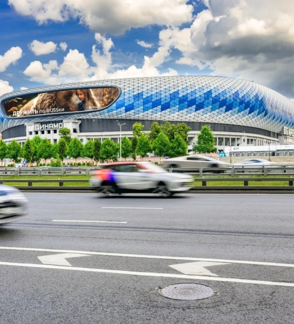 Sleek oval stadium with a blue diamond-pattern exterior beside a busy highway; blurred cars pass in the foreground.