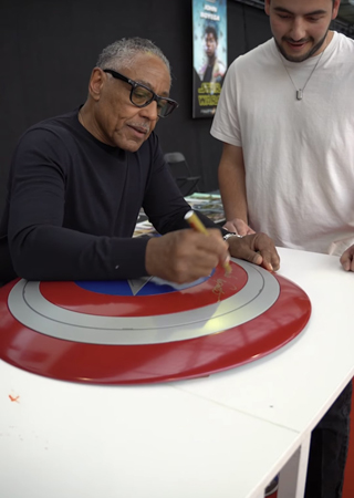 Джанкарло Эспозито подписывает щит Капитана Америки.