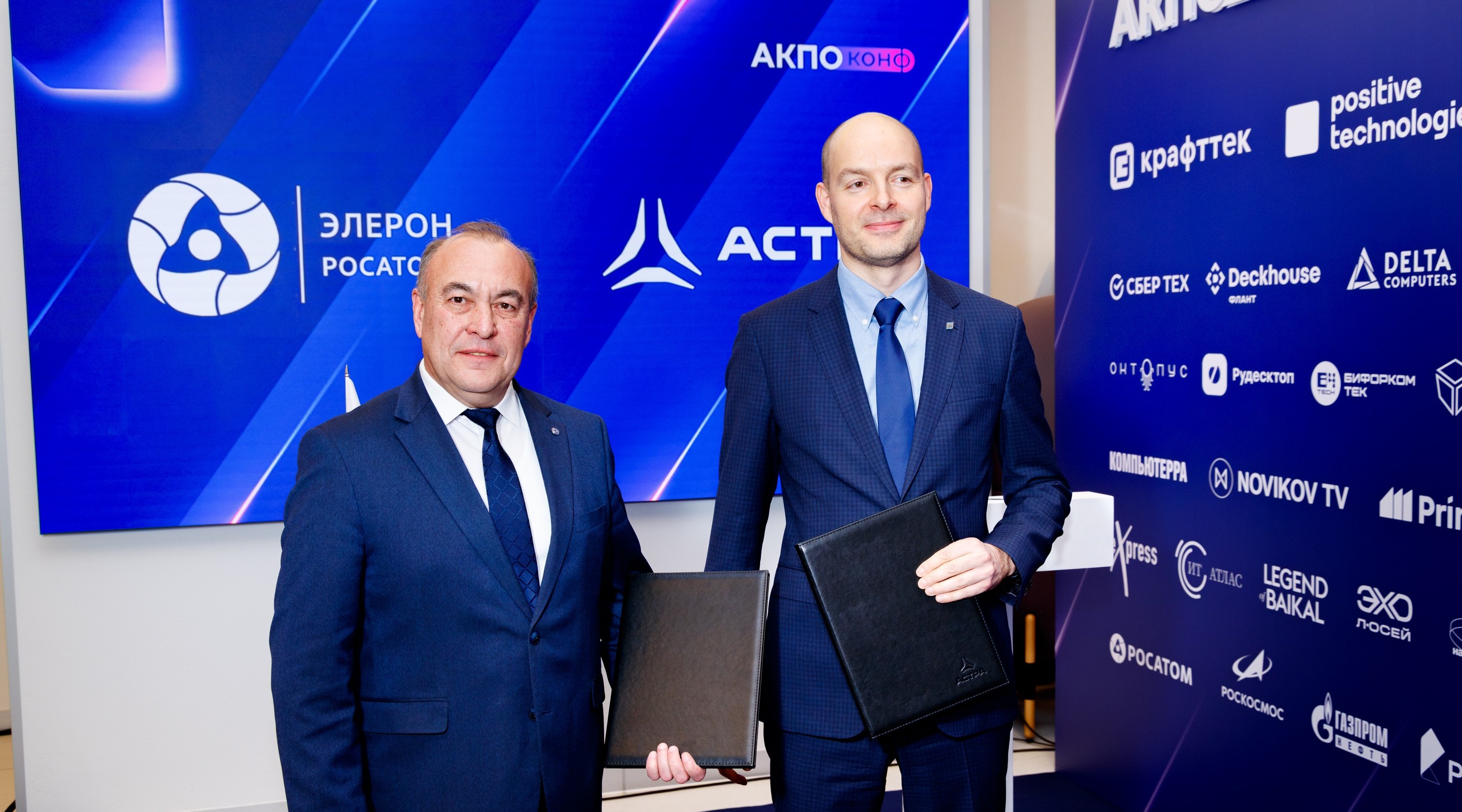 Two men in dark suits pose for a photo in front of a blue event backdrop, each holding a black folder.