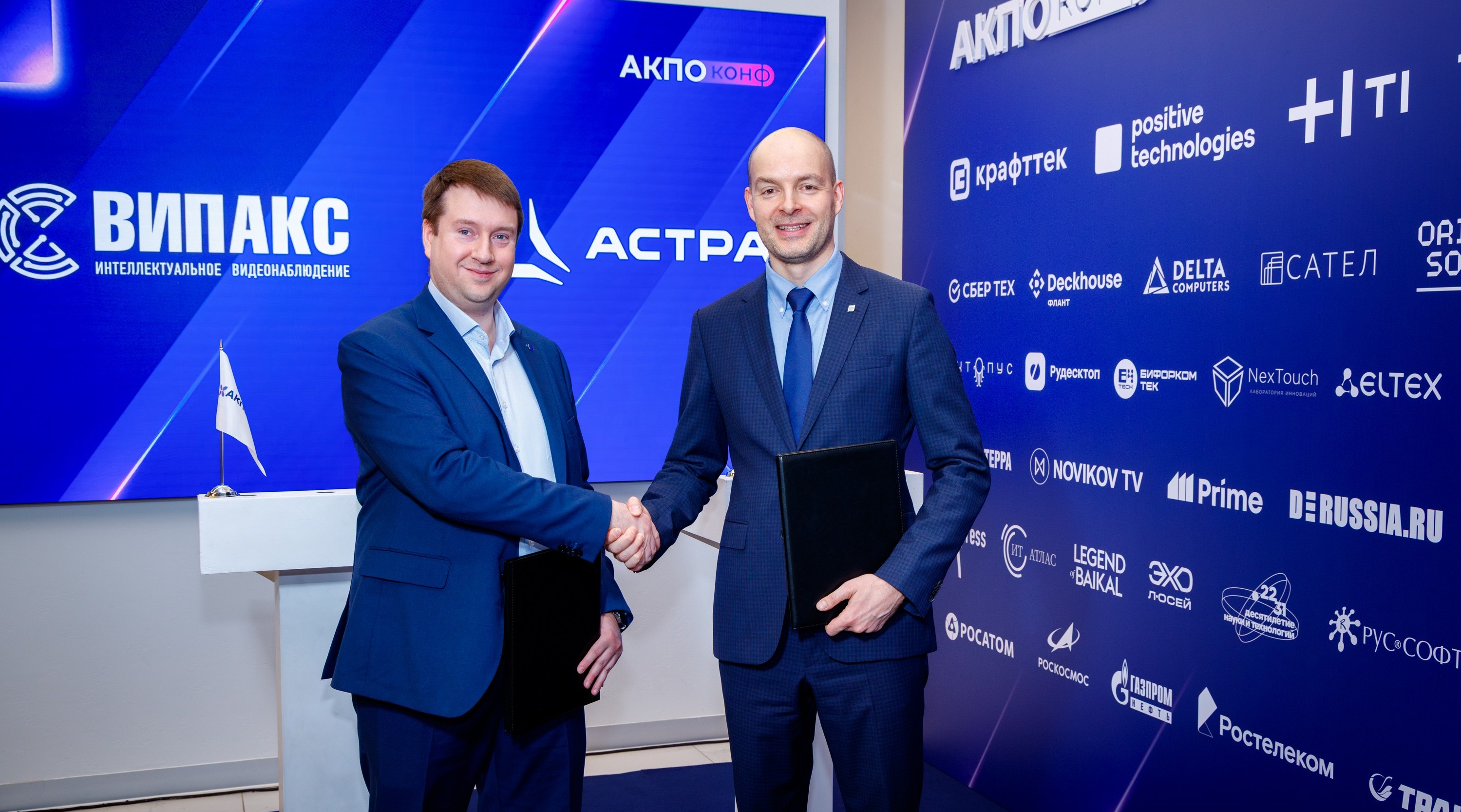 Two businessmen in suits shake hands at a conference booth with a blue sponsor backdrop.