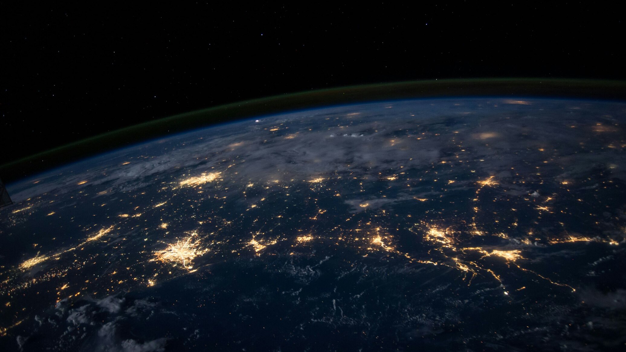 A view of Earth from space at night, with glowing city lights across the surface and a green aurora arc along the edge of the atmosphere.