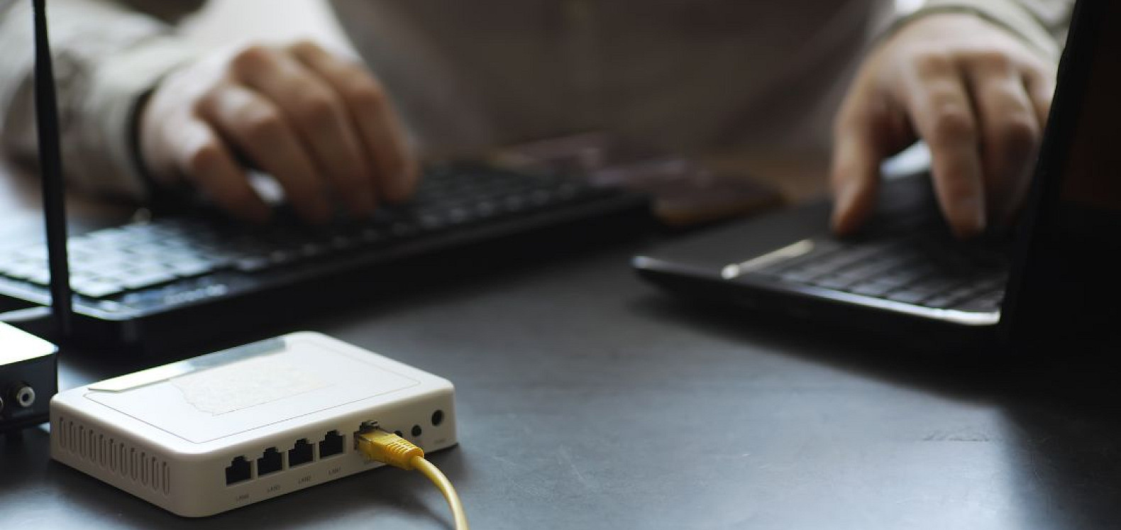 White network router with a yellow Ethernet cable plugged in on a dark desk, while two people type on laptops in the background.