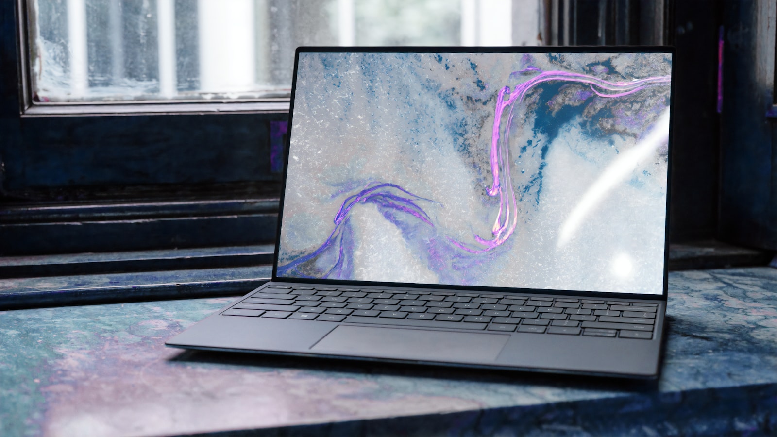 Laptop on a colorful, marbled desk with an abstract purple-pink wallpaper on the screen near a window.