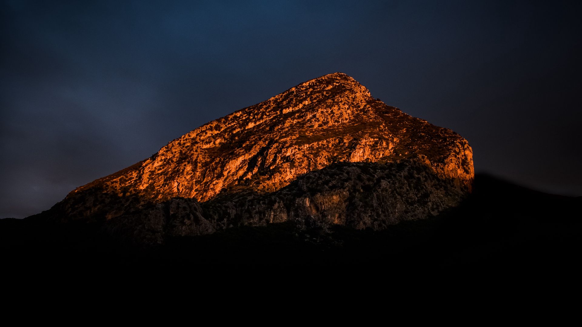 Rocky mountain peak illuminated in orange light against a dark blue twilight sky, with shadowy foreground.