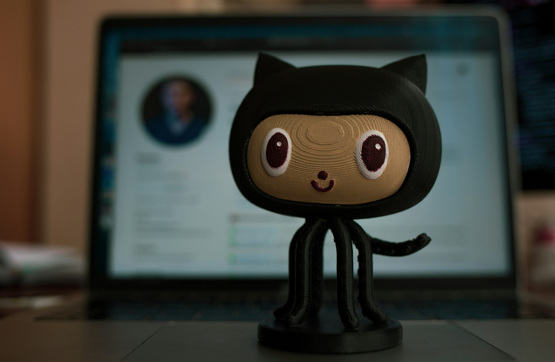 Close-up of a small cat-eared hooded plush on a desk in front of a blurred laptop screen.