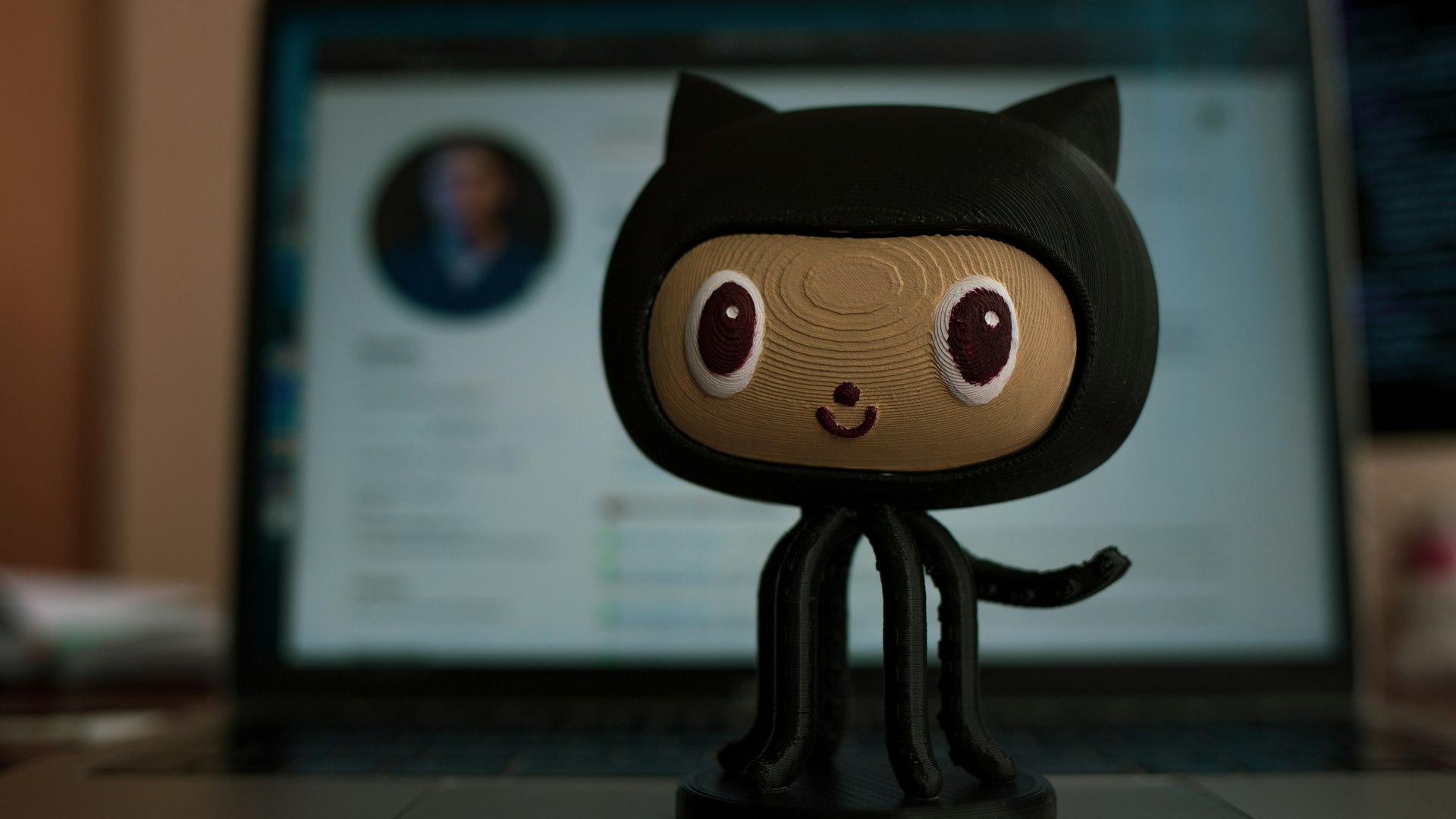 Close-up of a small plush figure wearing a black hoodie, with large eyes, on a desk in front of a blurred laptop screen in the background.