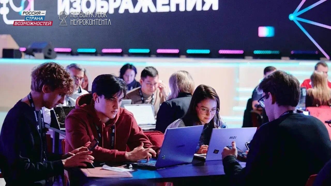 Group of young people working together at a round table with laptops in a conference setting, neon lights and a large screen behind them showing Russian text.
