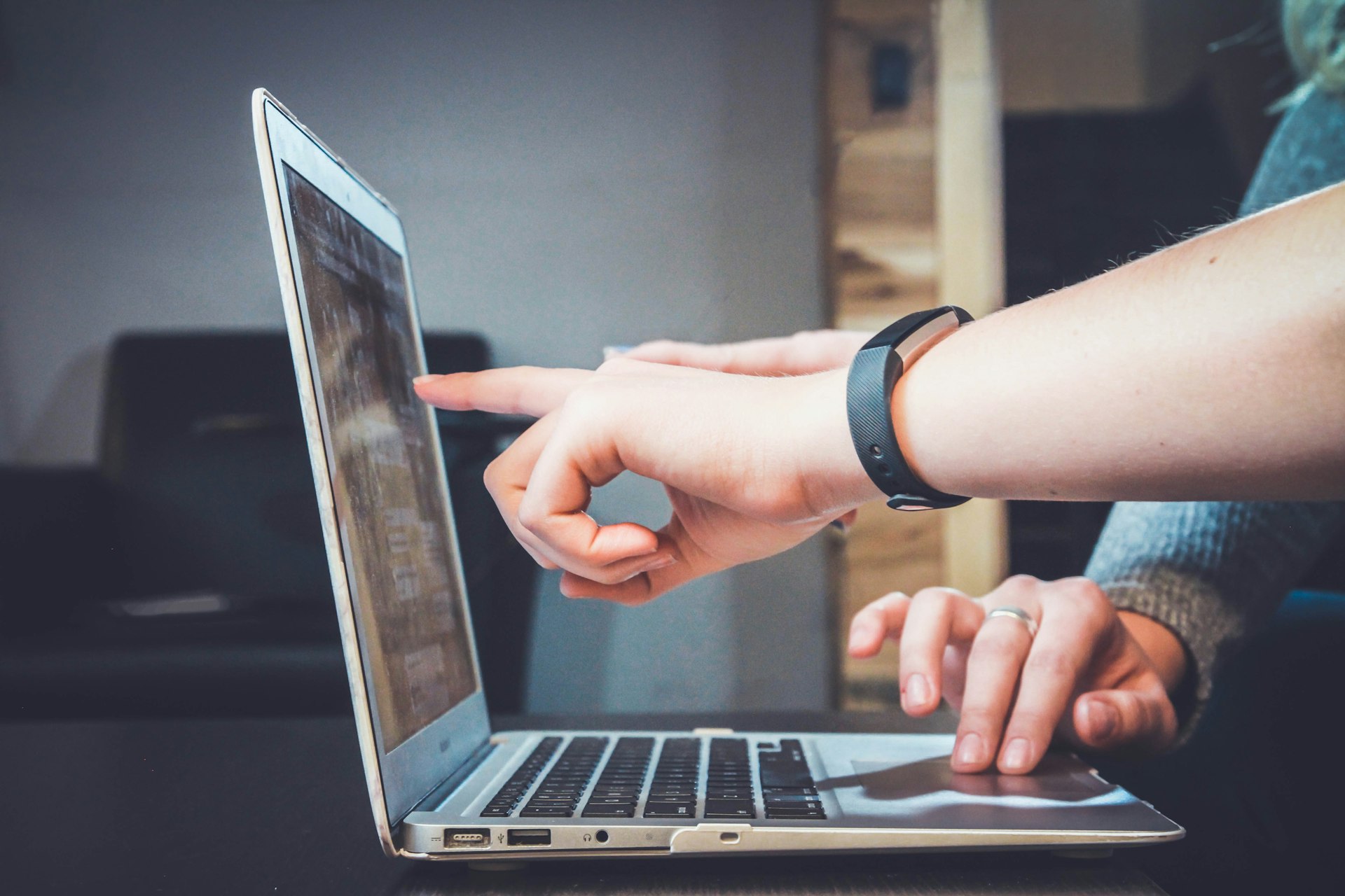 Two people collaborate at a laptop; one finger taps the screen while the other types her keyboard.
