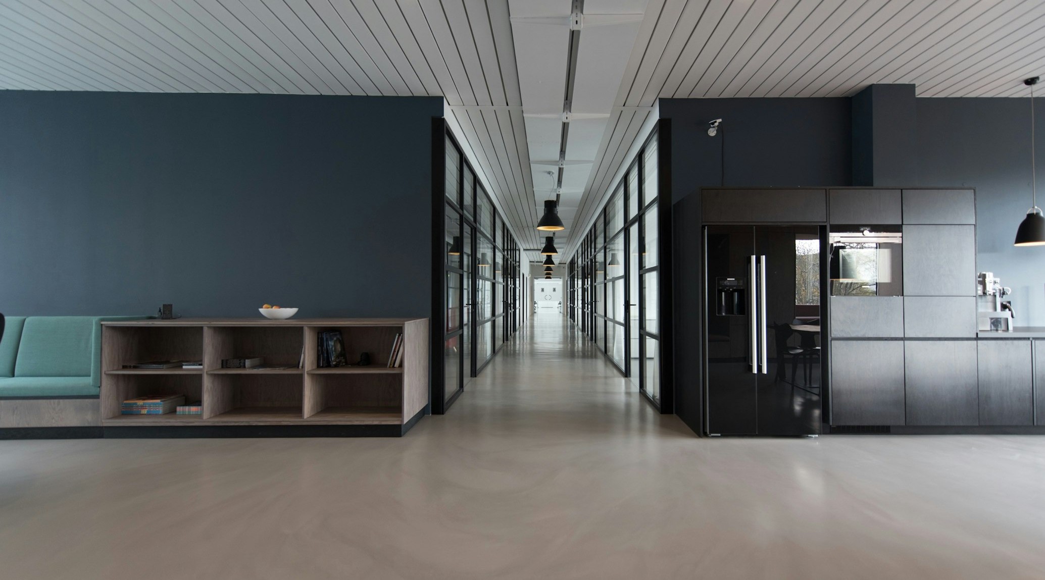 Modern office hallway with glass-walled rooms on both sides and a kitchenette area to the right, under hanging lights.