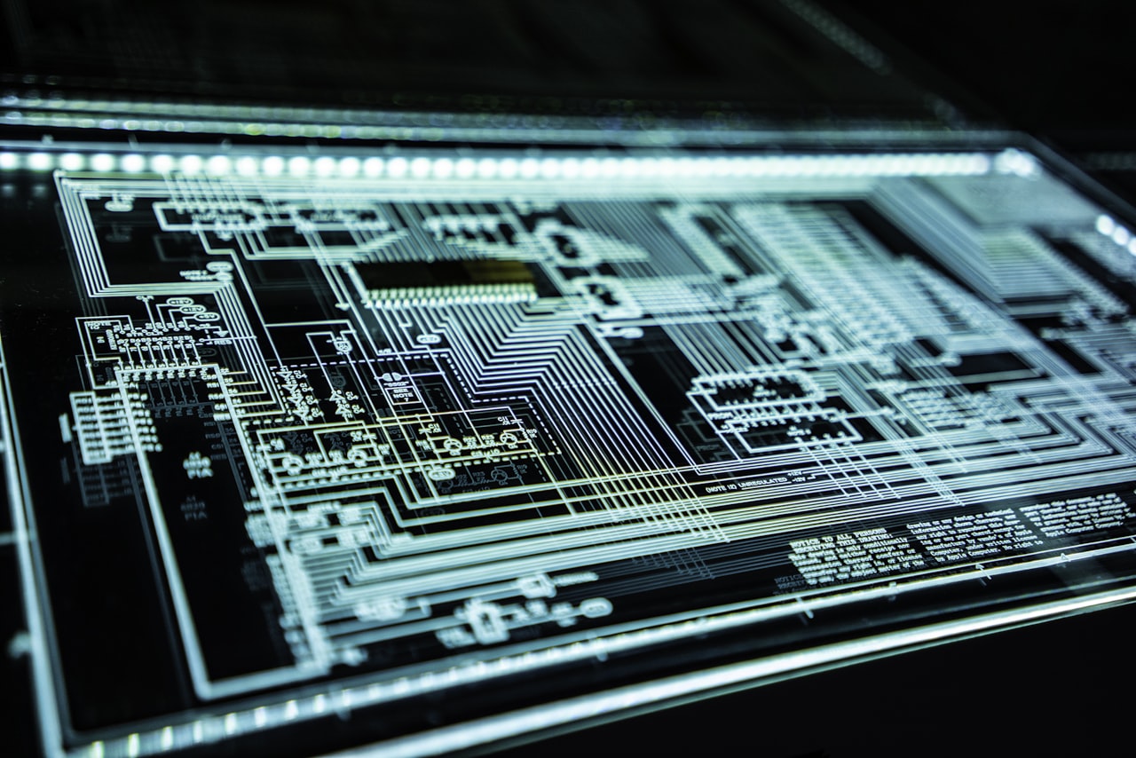 Close-up of a teal-glowing circuit board with intricate traces and microchips laid out in a dense PCB pattern.