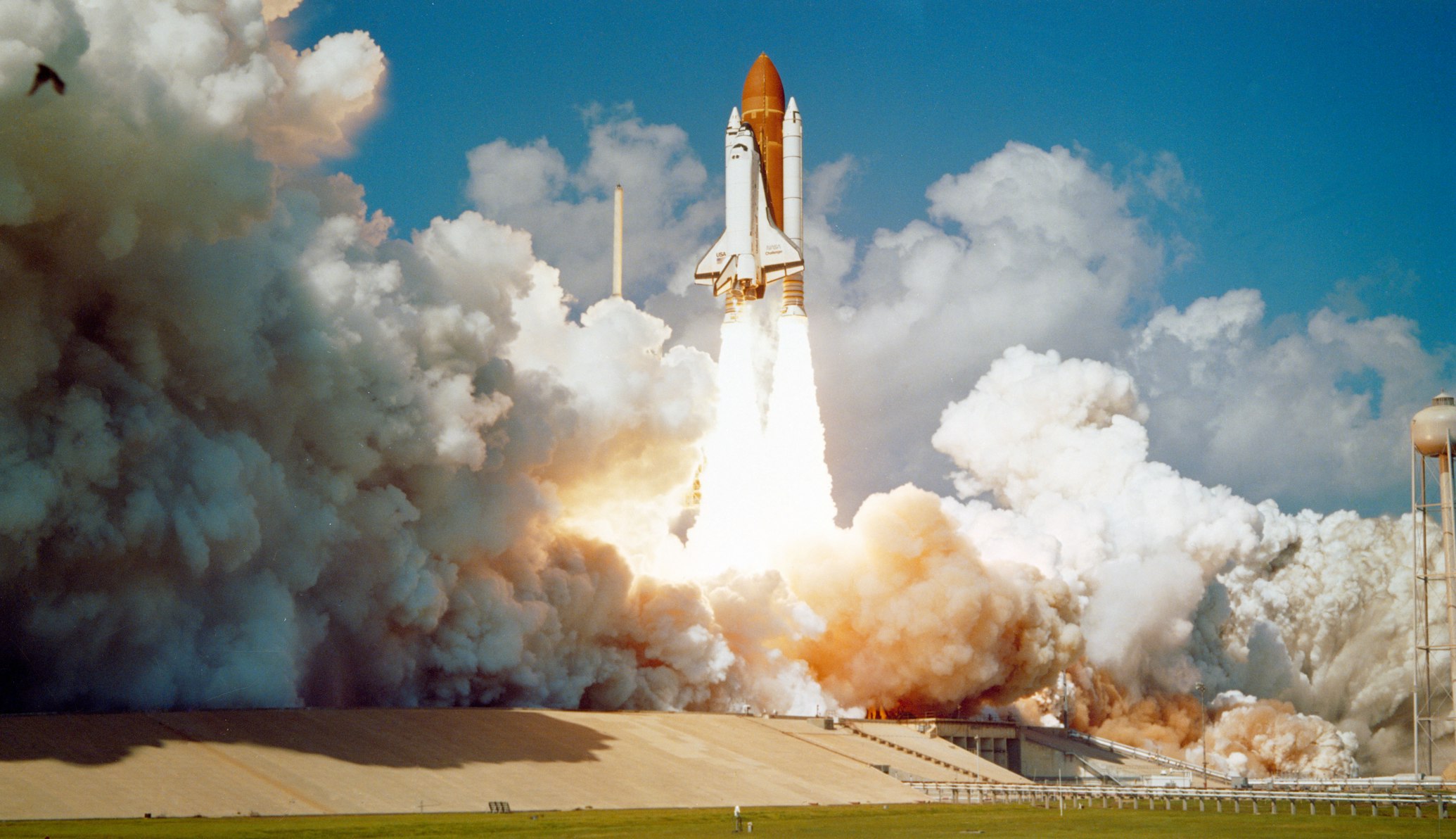 Space shuttle launches from a pad with orange external tank and white boosters, surrounded by thick smoke and flames.