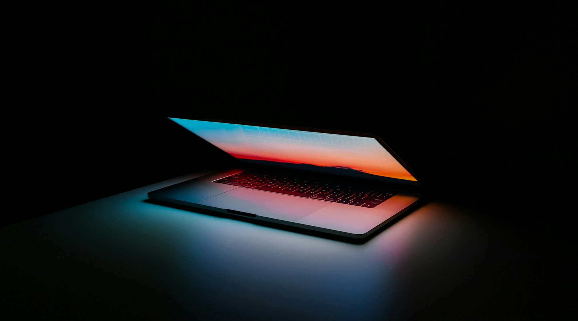 Laptop in a dark room with a colorful sunset gradient on the screen and a soft glow reflecting on the keyboard and desk.