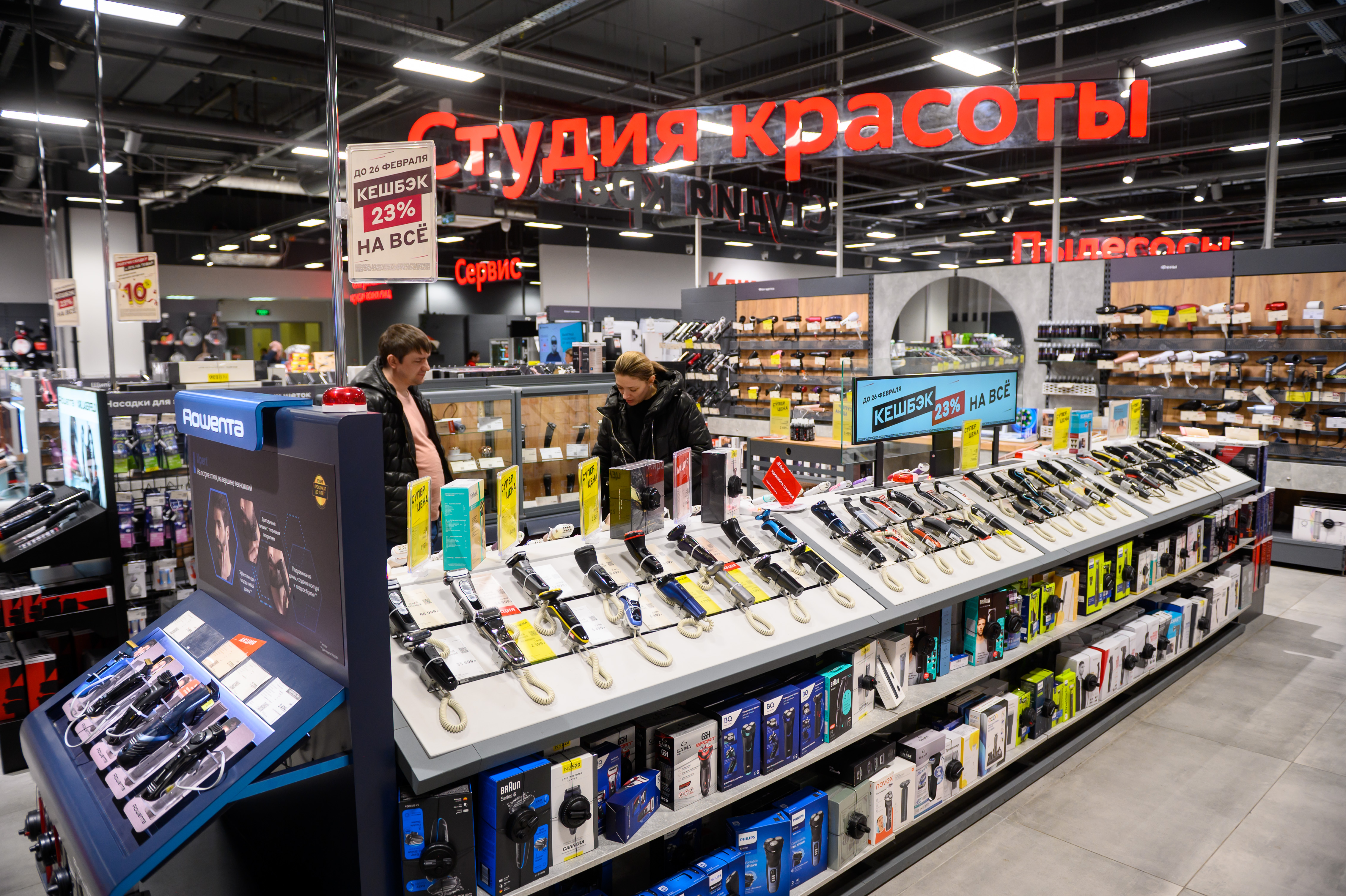 Store aisle in a electronics shop with rows of hair dryers and grooming devices on display; red Cyrillic signs overhead and two customers browsing nearby.