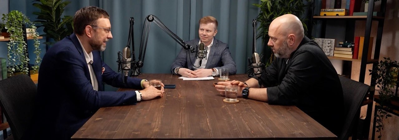 Three men sit around a wooden table in a podcast studio, speaking into large microphones with glasses in front of them.