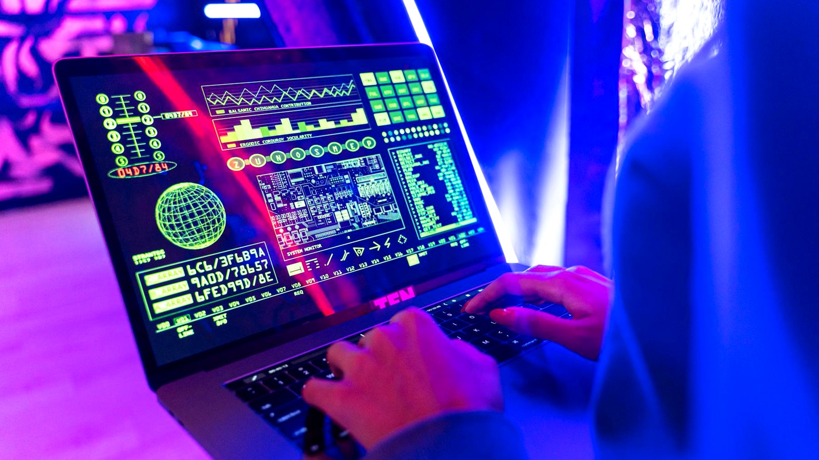 Person typing on a laptop with neon green and blue data dashboards and a glowing globe on screen in a dark, colorful setting.