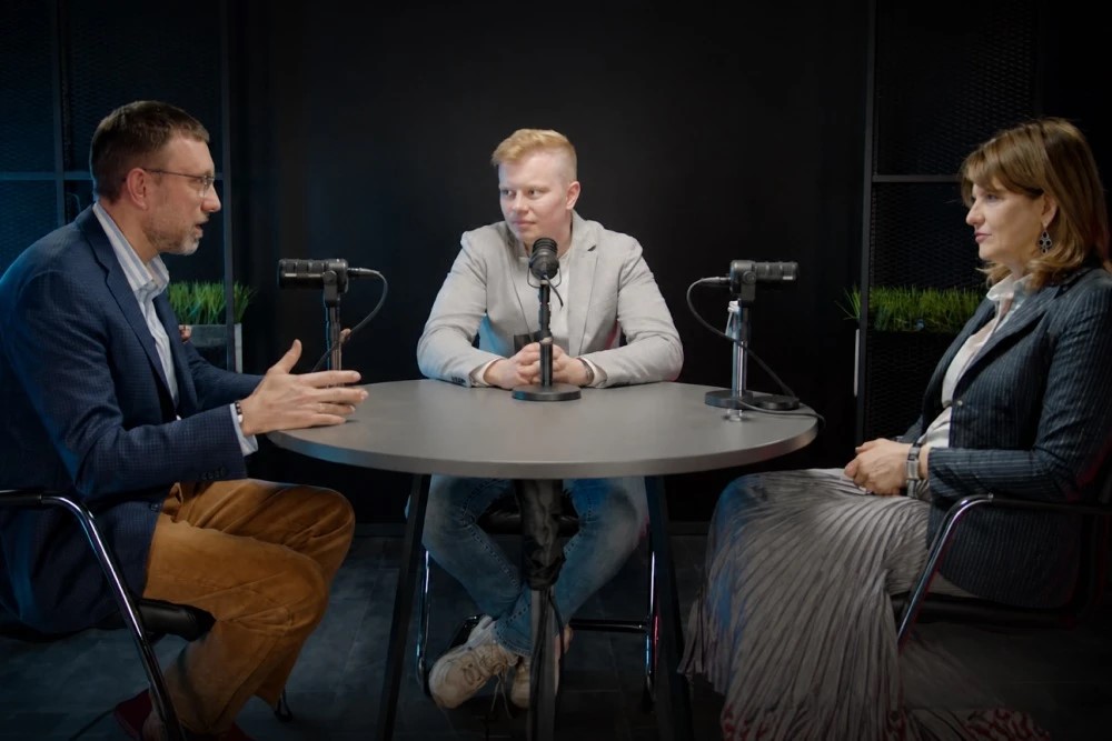 Three people sit around a round table in a podcast studio, each with a microphone, engaged in discussion.