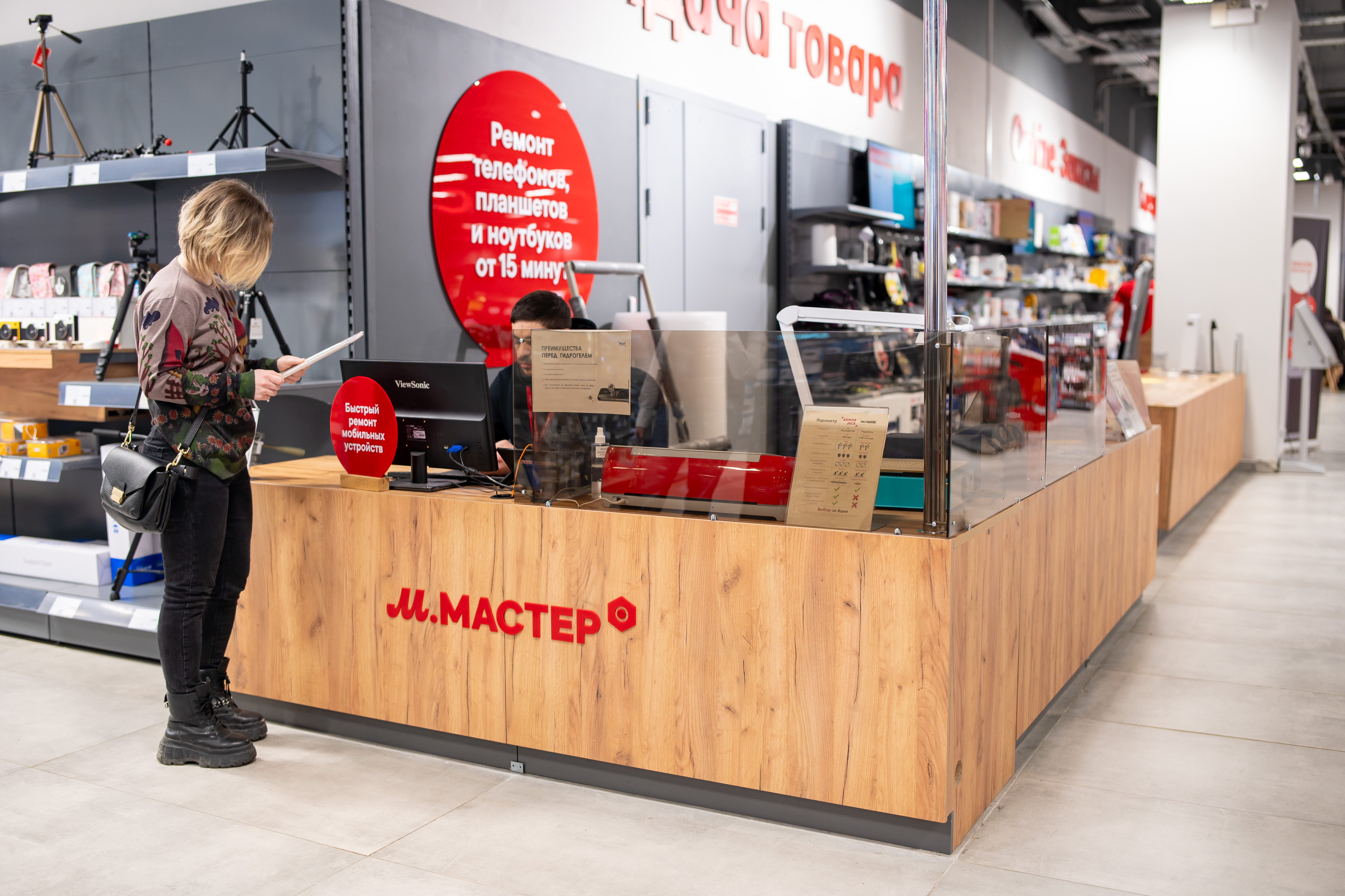 Female customer with a shoulder bag at a wooden checkout counter in an electronics store, looking at a document in her hand.