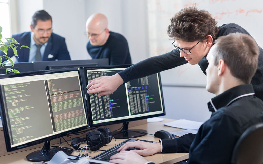 Three software developers collaborate at a desk with multiple monitors displaying code; one person points at the screen as others work.