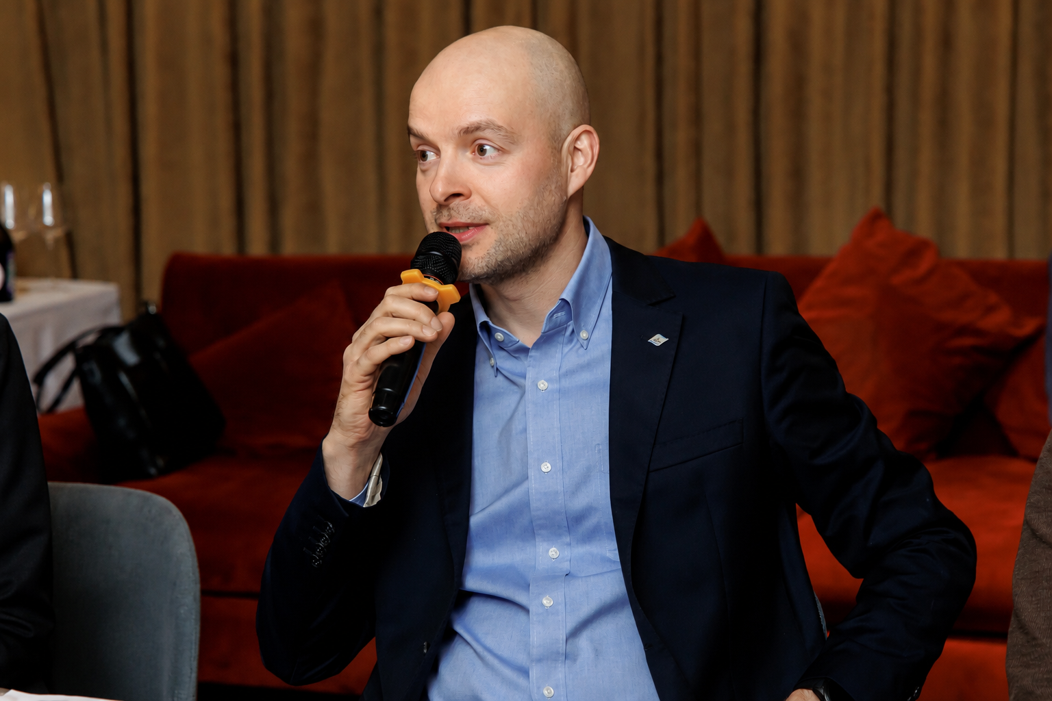 Man in a blue dress shirt and dark blazer speaks into a handheld microphone at an indoor event.