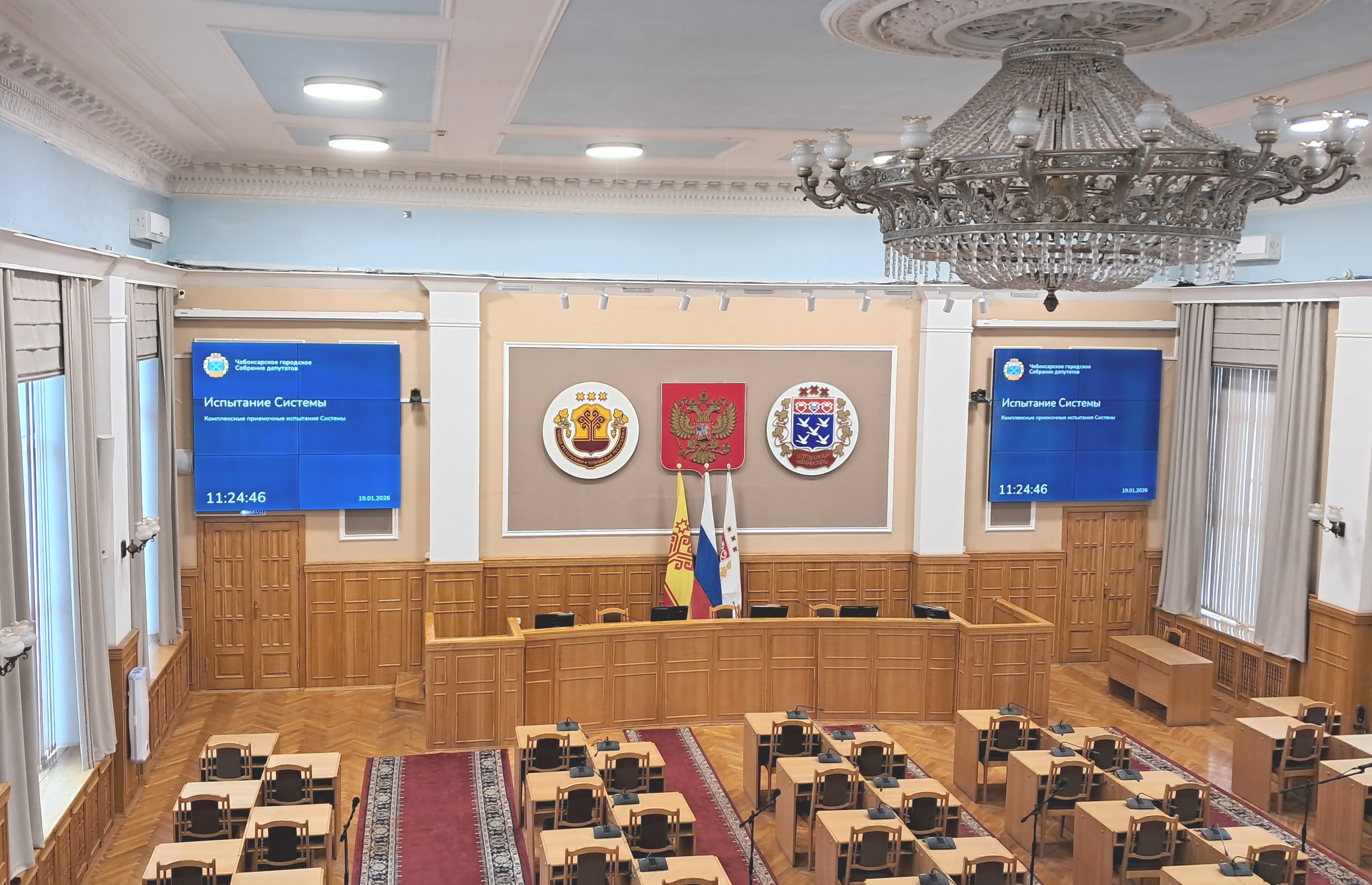 Large parliamentary chamber with an ornate chandelier, wooden desks, and three emblem plaques on the wall, plus blue presentation screens.