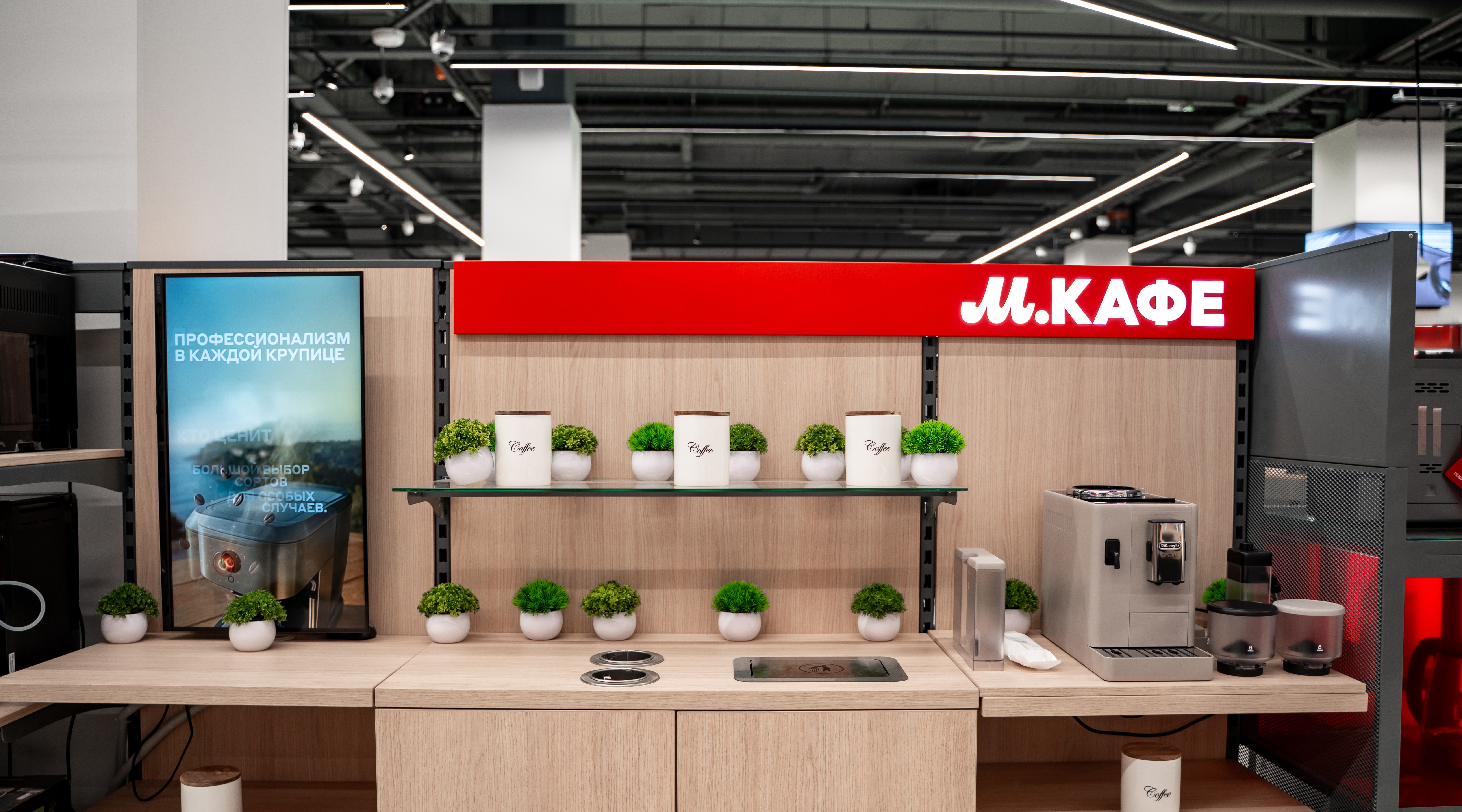 Coffee kiosk display with small potted plants and white canisters under a red М.КАФЕ sign in a retail space