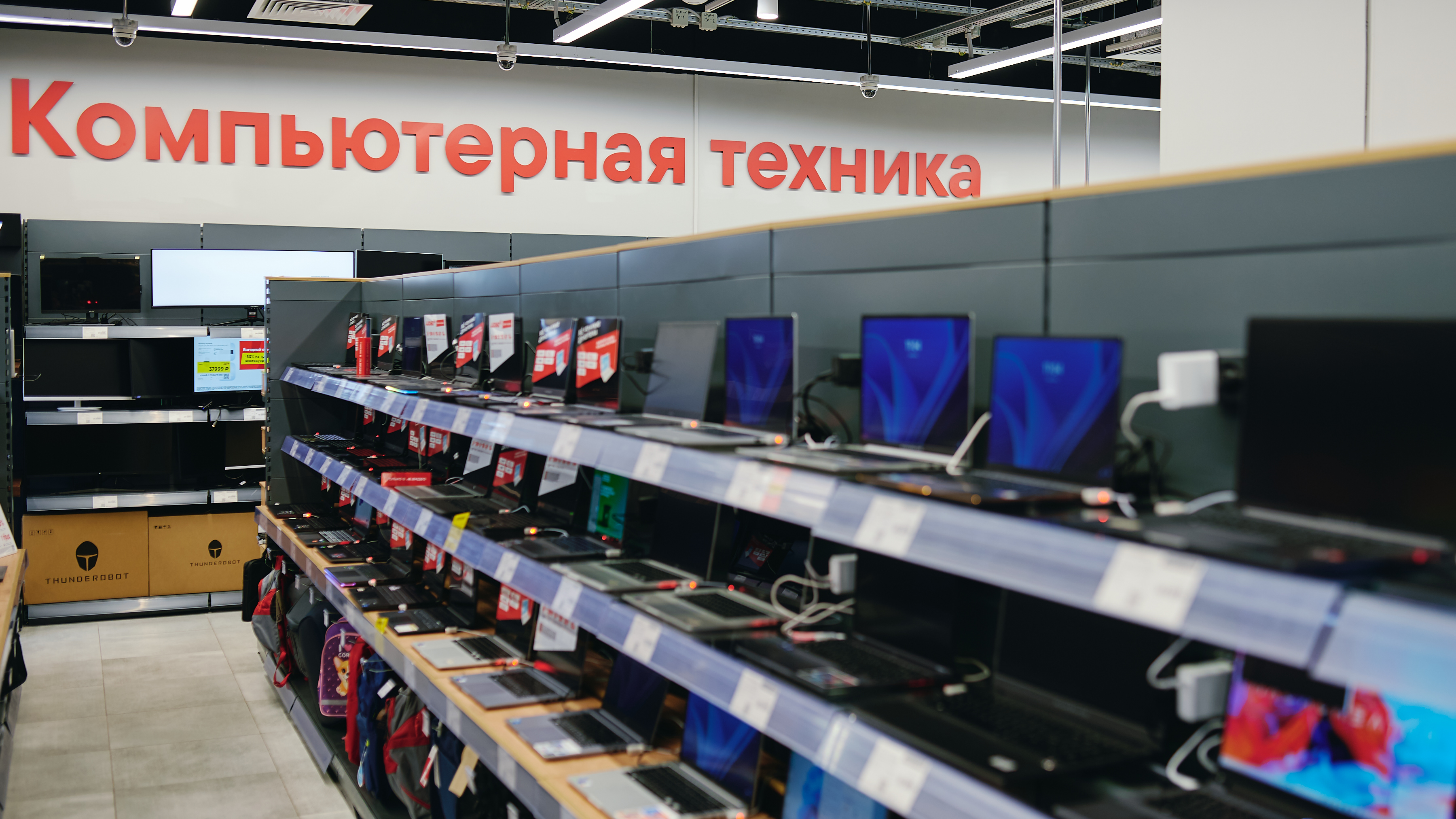 Row of laptops on display along shelves in a computer shop, with a large red Cyrillic sign reading Компьютерная техника above.