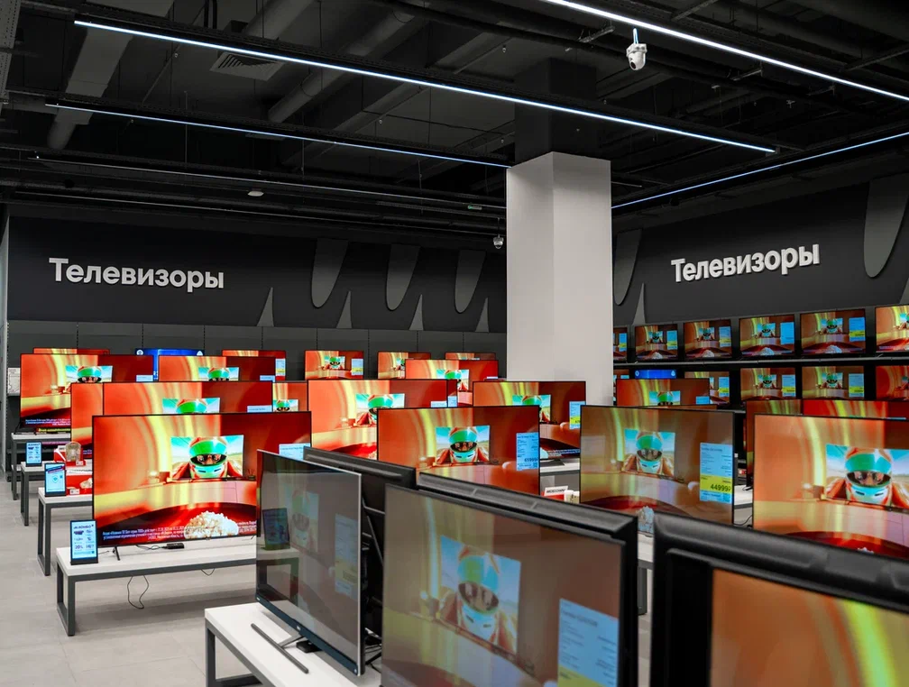 Showroom displaying numerous flat-screen TVs on stands under a dark ceiling with a Russian 'Телевизоры' sign in the background.