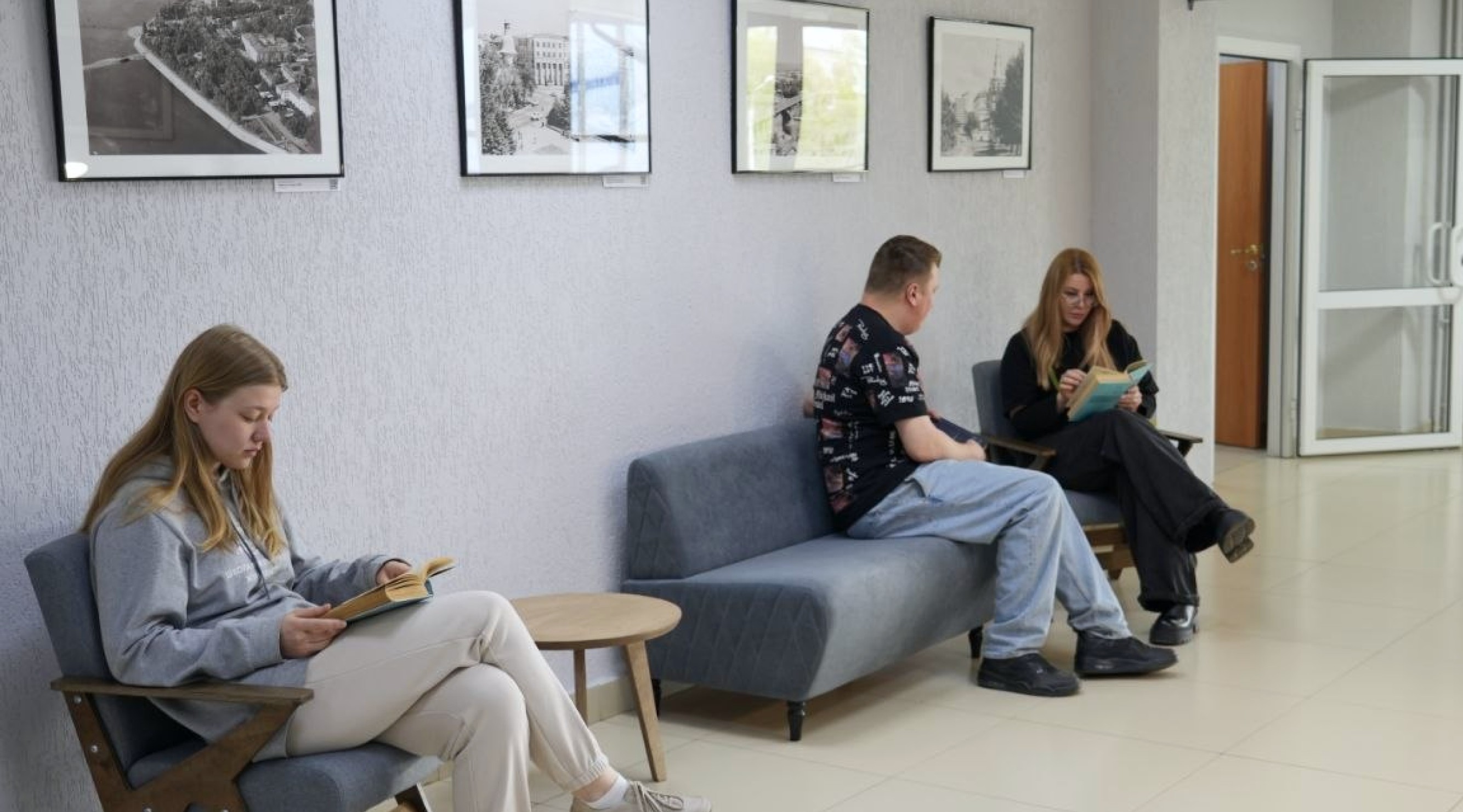 Three people sit on a grey couch area, each reading a book in a bright lobby with framed photos on the wall.