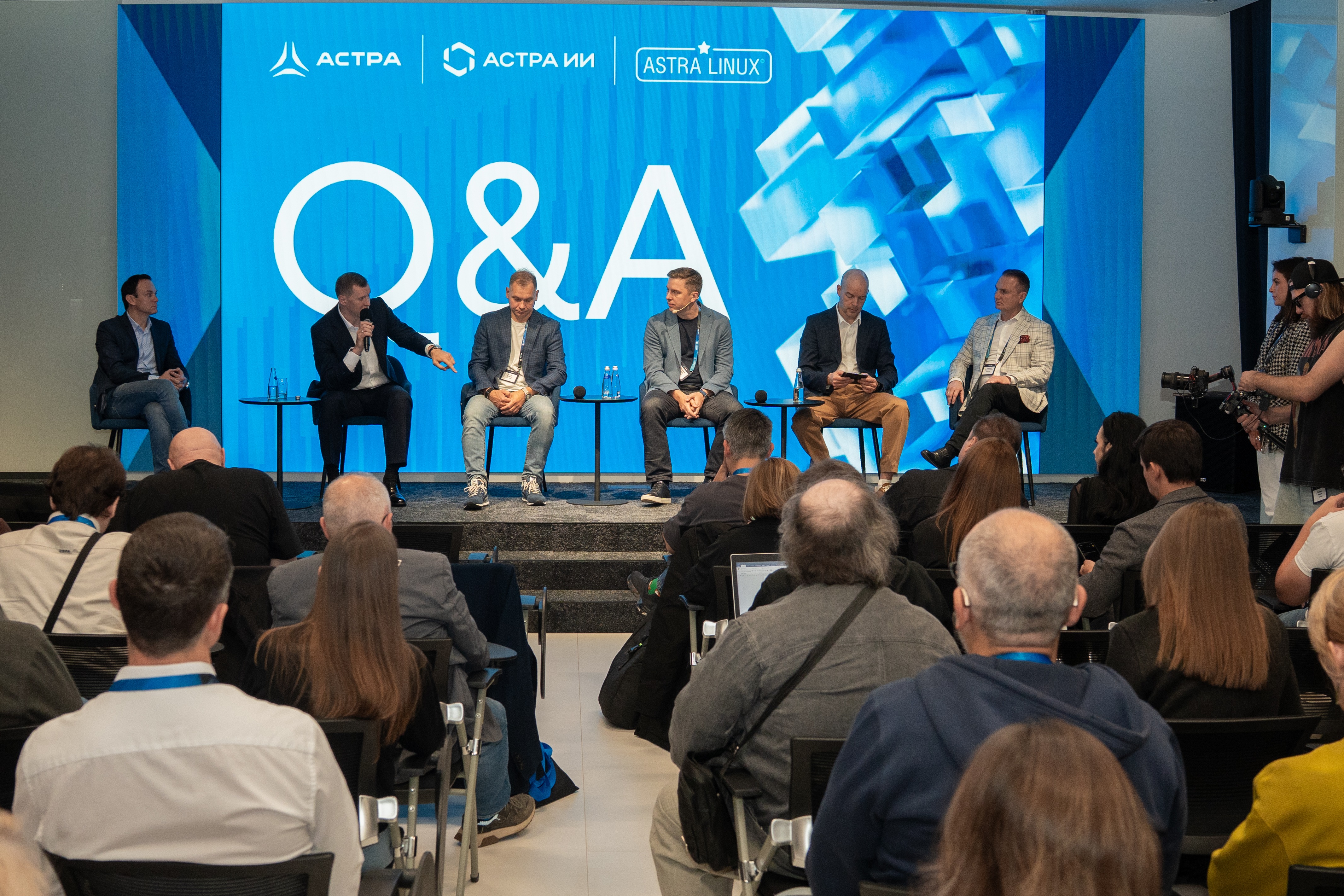 Six panelists seated on a stage during a Q&A session with a blue backdrop marked 'Q&A' behind them. A seated audience watches the discussion.