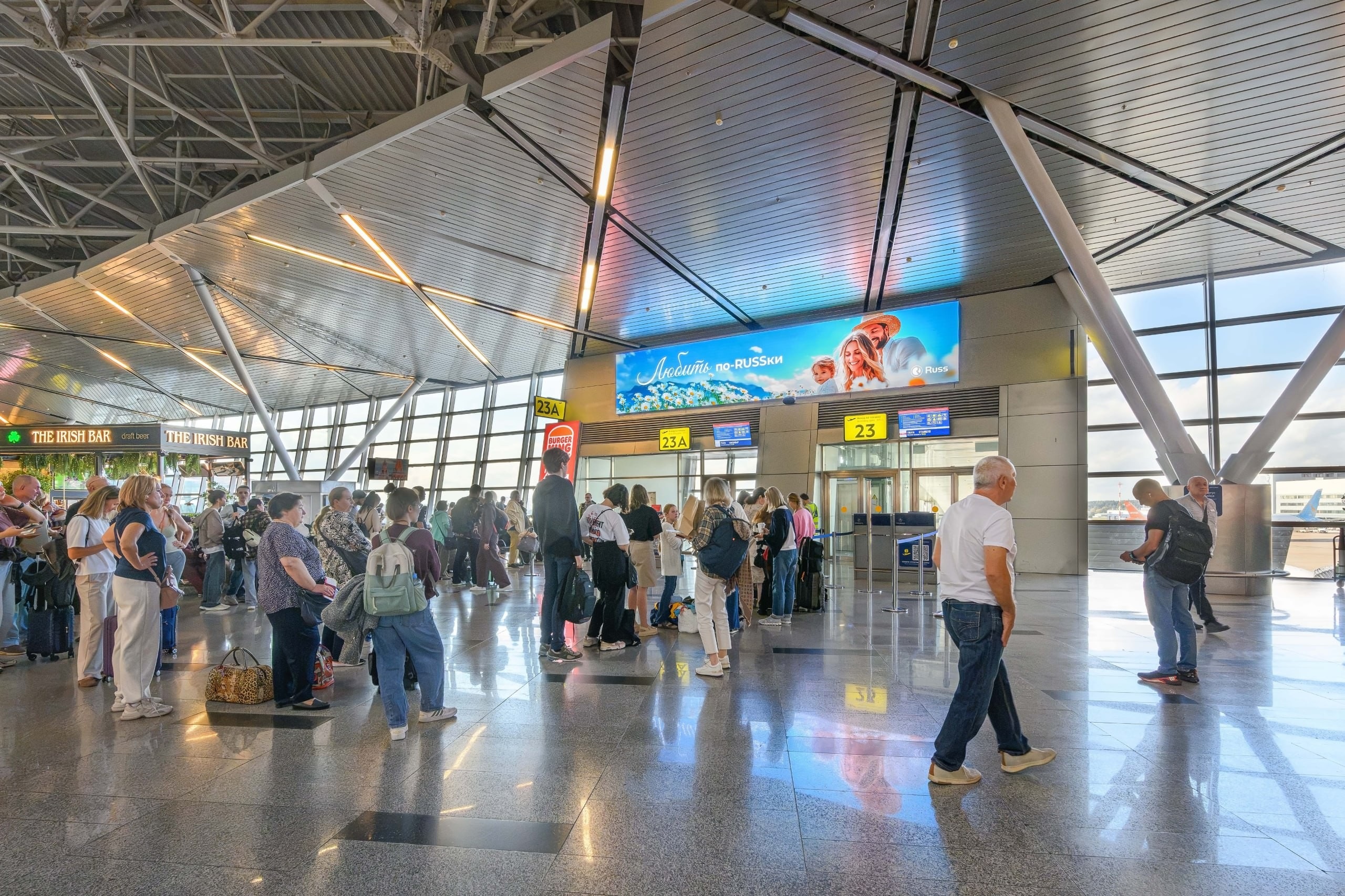 Crowded airport terminal with passengers waiting in line and luggage nearby, against large windows and modern architecture.