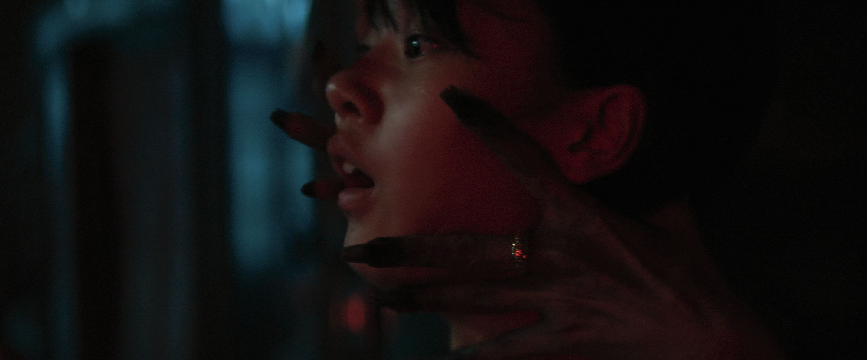 Close-up of a person in dark blue-red lighting, mouth slightly open as a finger rests near lips, wearing a ring on the finger and red nails visible in low light.
