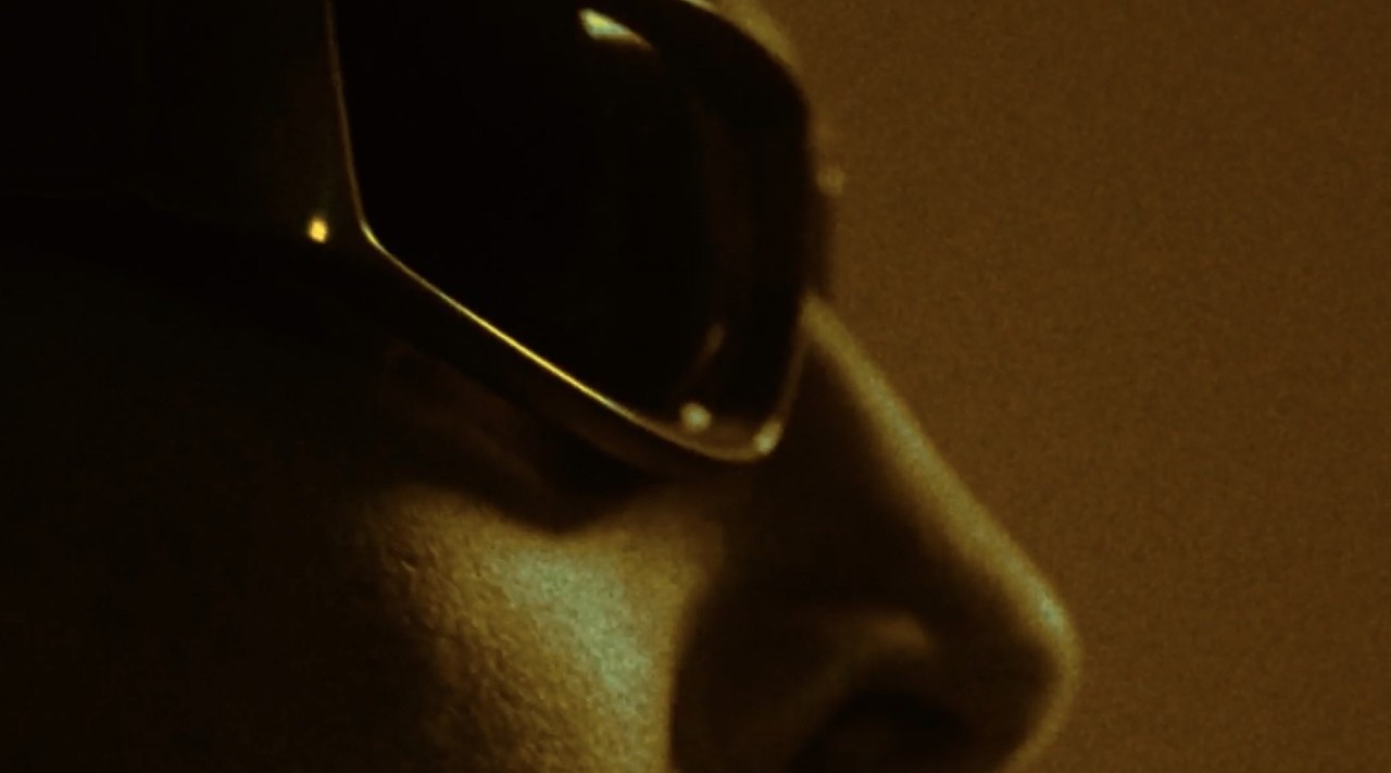 Close-up of a person wearing dark sunglasses in low light, showing the nose and cheek area with amber lighting.