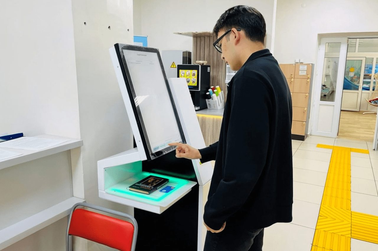 Man in a dark jacket uses a self-service touchscreen kiosk in a bright lobby area, touching the screen.